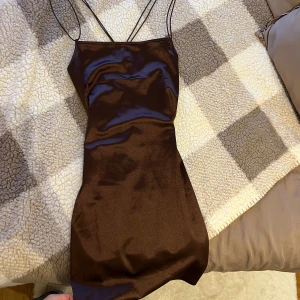 Brun klänning, S - Snygg brun klänning i satin med tunna axelband som korsas i ryggen. Klänningen är figursydd och har en rak, kort kjol. Från hm och i gott skick!