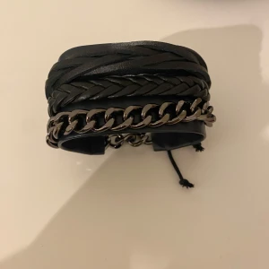 Svart läderarmband med kedjedetalj - Snyggt svart armband i läder med flätade detaljer och en silverfärgad kedja. Justerbar passform med snörning för en cool och modern look.Helt ny .Aldrig använd 