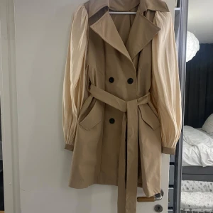 Beige trenchcoat/klänning med bälte - Snygg beige trenchcoat/klänning med knytbälte i midjan och dubbelknäppning framtill. Den har långa ärmar med en lätt puffig design och klassisk krage. Perfekt för vår och höst.