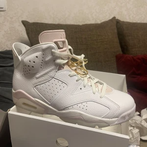 Nike Air Jordans i vitt och rosa - Säljer ett par stilrena Nike Air Jordans i vitt med rosa detaljer. Skorna har snörning och är tillverkade i syntetmaterial. Perfekta för den som vill ha en klassisk och sportig look.