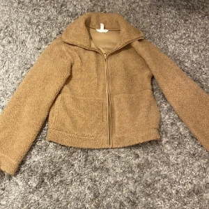 Beige teddyjacka med dragkedja - Mysig och varm beige teddyjacka perfekt för kyliga dagar. Jackan har en dragkedja framtill och en hög krage som håller dig varm. Den är långärmad och har en avslappnad passform, vilket gör den idealisk för både vardag och utflykter.