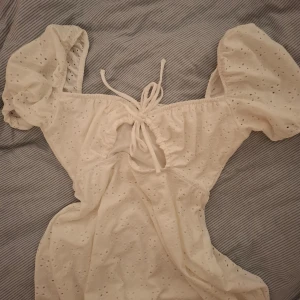 Vit klänning från H&M Divided - Charmig vit klänning från H&M Divided med puffiga kortärmar och knytning framtill. Klänningen har ett broderat mönster och cut-out