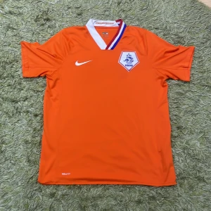 Nederländerna/Holland fotbollströja från Nike - Säljer en orange fotbollströja från Nike med KNVB-logga på bröstet. Tröjan har en vit och röd krage och är kortärmad. Perfekt för fotbollsfans som vill visa sitt stöd.