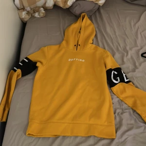 Gul hoodie från Jack & Jones - Säljer en snygg gul hoodie från Jack & Jones med texten 'DEFYING' på bröstet. Tröjan har svarta detaljer på ärmarna och en bekväm huva. Perfekt för en avslappnad stil.