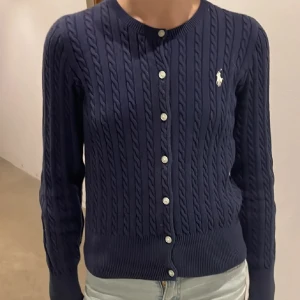 Mörkblå kofta från Ralph Lauren - Snygg mörkblå kofta från Ralph Lauren med kabelstickat mönster och knappar framtill. Den har långa ärmar och en liten logga på bröstet. Storlek M i barn storlek! Bilder av gamla ägare 