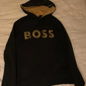 Svart hoodie från Boss - Snygg svart hoodie från Boss med stort logotryck på framsidan. Hoodien har en justerbar huva med snören och en kontrasterande beige insida. Perfekt för en avslappnad stil.