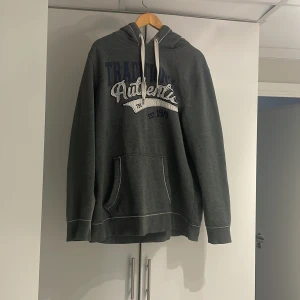 Grå hoodie från Livergy - Säljer en grå hoodie från Livergy med texten 'Traditional Authentic' på framsidan. Den har en stor ficka framtill och justerbar huva med vita snören. Perfekt för en avslappnad stil.