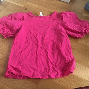 Rosa topp med puffärmar - Säljer en snygg rosa topp med puffiga kortärmar. Perfekt för att ge en färgklick till din garderob. Toppen har en rund halsringning och är tillverkad i ett mjukt material.