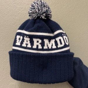 Mörkblå mössa med vit ”VÄRMDÖ” text  - Snygg mörkblå mössa med vit text 'Värmdö' och en matchande tofs på toppen. Perfekt för kalla dagar.