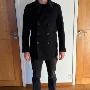 Svart skepparkavaj John Henric  - Snygg svart skepparkavaj med dubbelknäppning och klassisk krage. Jackan har långa ärmar och en stilren design som passar perfekt för kyligare dagar. Den har även en dekorativ knappdetalj baktill.