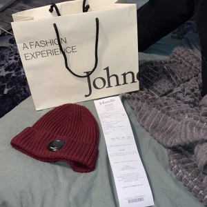 Vinröd stickad mössa från C.P. Company - Snygg vinröd stickad mössa från C.P. Company med ribbad design och en rund svart logotyp framtill. Perfekt för att hålla värmen med stil. Köpt ifrån jhonells. Kvitto medföljer 