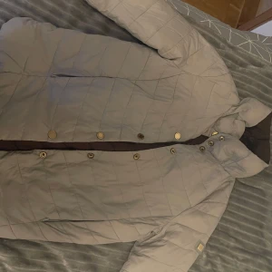 Vändbar quiltad jacka från pedro del Hierro  - Säljer en snygg vändbar quiltad jacka från RRL. Ena sidan är beige och den andra är brun, vilket ger dig två stilar i en. Jackan har knappar och en hög krage för extra värme. Perfekt för kyliga dagar!Du kan ha på dig jackan åt båda hållen. Storlek är S och M 