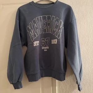 Blå sweatshirt från Gina Tricot - Säljer en blå sweatshirt från Gina Tricot med texten 'Maverick 67 Sports USA' tryckt på framsidan. Tröjan har långa ärmar och ribbade muddar vid ärmslut och nederkant. Perfekt för en avslappnad stil.