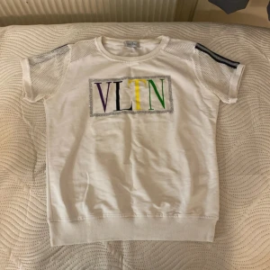 Vit topp från Valentino med VLTN-tryck - Snygg vit topp från Valentino med färgglatt VLTN-tryck på framsidan. Toppen har korta ärmar med nätmaterial och svarta ränder. Perfekt för en trendig look!