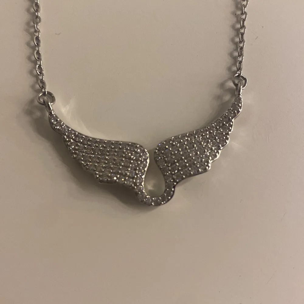 Elegant silverfärgat halsband med en unik vingdesign. Kedjan är tunn och smidig med ett justerbart lås. Vingarna är dekorerade med små glittrande stenar som ger en lyxig känsla. Perfekt för att lägga till en touch av glamour till din outfit.. Asusteet.