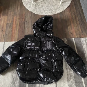 Svart glansig pufferjacka med huva - Säljer en svart glansig pufferjacka med huva. Jackan har en cool textdetalj på bröstet och ryggen. Perfekt för att hålla värmen med stil. Den har en dragkedja framtill och är ärmlös, vilket gör den mångsidig för olika väder. Skick 10/10 riktigt fet och passar perfekt. Storlek S priset är inte hugget i sten!