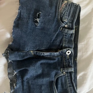 Blå jeansshorts med slitningar - Säljer ett par coola blå jeansshorts från Disel. De har slitna detaljer och dragkedjor på bakfickorna. Perfekta för en avslappnad stil. Midjan är låg och de har en knapp och dragkedja framtill.