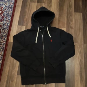 Svart hoodie från Ralph Lauren - Säljer en stilren svart hoodie från Ralph Lauren med dragkedja och en liten röd logotyp på bröstet. Tröjan har långa ärmar och en justerbar huva med vita snören. Perfekt för en avslappnad look.(XS-S) passar runt 164-170.