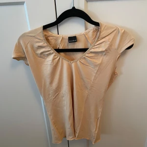 Beige kortärmad topp från Bruuns Bazaar - Säljer en stilren beige topp från Bruuns Bazaar. Den har en v-ringad design med korta ärmar och fina guldglittriga detaljer vid halsringningen. Perfekt för en elegant och avslappnad look. Skulle säga att den är i XS men passar även S. Köpare står för frakten 🩷