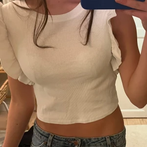 Vit ribbad topp från Zara - Säljer en vit ribbad topp från Zara med korta ärmar och volangdetaljer. Toppen är croppad och har en tight passform, perfekt för sommarens varma dagar. Passar bra till jeans eller kjol för en avslappnad look.