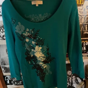 Grön/blå tröja från Signature Experience - Snygg topp från Signature Experience med ett unikt blommönster i svart och vitt på framsidan. Tröjan har trekvartsärmar och en rund halsringning. Perfekt för att ge en färgklick till din garderob!