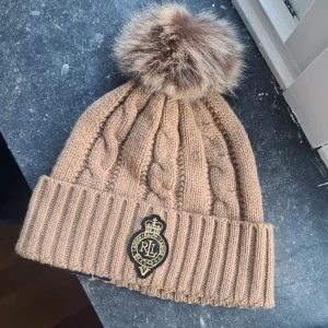 Beige stickad mössa från Ralph Lauren - Snygg beige stickad mössa från Ralph Lauren med en fluffig pälsboll på toppen. Mössan har ett elegant kabelstickat mönster och en broderad logotyp framtill. Perfekt för att hålla dig varm och stilren under de kallare månaderna.