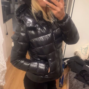 Svart pufferjacka från Moncler - Snygg svart pufferjacka från Moncler med glansigt material och huva. Jackan har en dragkedja framtill och långa ärmar. Jackan är i modellen bady och säljer den så billigt eftersom den har en liten defekt på framsidan och har inte kvar äktahetsbeviset, kontakta för fler bilder, pris kan diskuteras 