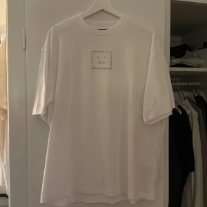 Vit t-shirt från Acne Studios - Säljer en stilren vit t-shirt från Acne Studios med ett unikt ansikte av pärlor på bröstet. T-shirten har en normal passform och korta ärmar, perfekt för en avslappnad look. NYPRIS: 2499:-