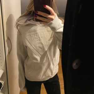 Grå hoodie från Guess - Säljer en stilren grå hoodie från Guess med en stor logga på bröstet. Tröjan har en klassisk känguruficka och en bekväm huva. Perfekt för en avslappnad look.