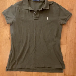 Olivgrön pikétröja från Ralph Lauren - Säljer en olivgrön pikétröja från Ralph Lauren med klassisk krage och korta ärmar. Tröjan har en broderad logotyp på bröstet och är i modellen 'The Skinny Polo'.  Den har ett litet hål på baksidan under kragen som knappt märks av. Hör av dig vi minsta lilla fundering. Passar dig som e 155-165