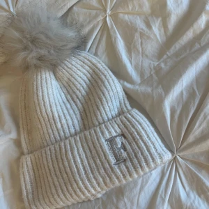 Vit ribbad mössa med pälsboll - Säljer en stilren vit ribbad mössa med en fluffig pälsboll på toppen. Mössan har en broderad bokstav 'F' framtill. Perfekt för kyliga dagar. Köpte den från Primark. Aldrig använd endast testad.