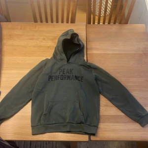 Grön hoodie från Peak Performance - Säljer en grön hoodie från Peak Performance med svart text på framsidan. Den har en klassisk design med långa ärmar och en rymlig huva. Perfekt för en avslappnad stil.