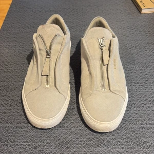 Gråa sneakers med dragkedja från Axel Arigato - Snygga gråa sneakers från Axel Arigato i mocka med dragkedja framtill. Skorna har en stilren design med en platt sula och är perfekta för en avslappnad look. Märkesnamnet är diskret präglat på sidan. Box + dustbag tillkommer allt og!