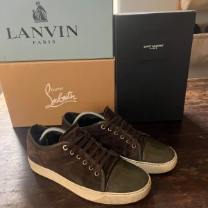 Bruna sneakers från Lanvin - Snygga bruna sneakers från Lanvin i mocka med snörning och en glansig tåhätta. Perfekta för en stilren look med en touch av lyx. Skorna har en vit sula som ger en fräsch kontrast till det mörkbruna materialet.