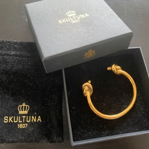 Guldpläterat armband från Skultuna - Elegant armband från Skultuna i guldpläterat 316L stål. Designen har detaljerade ändar som liknar djurhuvuden, vilket ger en unik touch. Levereras i en stilren svart ask med Skultunas logotyp. 