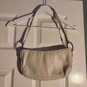 Beige axelväska från Carin Wester - Snygg beige axelväska från Carin Wester i ett stilrent och minimalistiskt design. Väskan har en praktisk dragkedja och är perfekt för att bära dina nödvändigheter. Den är gjord i ett mjukt material som ger en lyxig känsla.