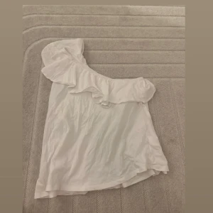 Vit oneshoulder topp - Säljer en söt vit oneshoulder topp med volangdetaljer. 