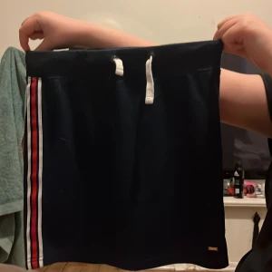 Mörkblå kjol med ränder - Snygg mörkblå kjol med vita och röda ränder längs sidorna. Kjolen har en bekväm resårmidja med vit dragsko för justerbar passform. Perfekt för en sportig look.