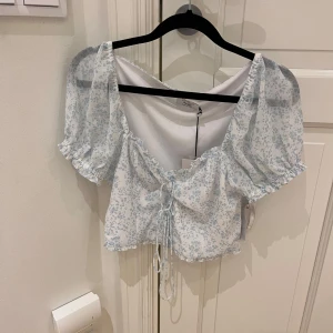 Blommigt sett  - Säljer en söt blommig blus från NA-KD i storlek S. Blusen har korta puffärmar och en knytning framtill. Den är vit med ljusblå blommönster och har en volangdetalj nedtill. Tillsammans med den matchande kjolen blir den perfekt för vår och sommar!