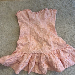 Rosa axelbandslös klänning - Charmig rosa axelbandslös klänning med volangdetaljer. Perfekt för sommarens festligheter. Den har en vacker struktur och en feminin siluett.