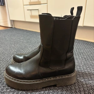 Svarta boots med dragkedja - Säljer ett par svarta boots i skinn med dragkedja på sidan. De har en robust sula och är perfekta för höst och vinter. Skorna har en klassisk design med rund tå och är lätta att matcha med olika outfits.