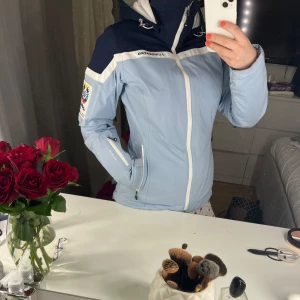 Ljusblå skidjacka från J. Lindeberg - Säljer en skitsnygg ljusblå skidjacka från J. Lindeberg med GORE-TEX-material. Jackan har en dragkedja framtill, justerbar huva och flera praktiska fickor. Perfekt för vintersport och håller dig varm och torr i backen. Den har snygga detaljer och en sportig look.