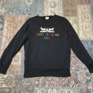 Svart tröja från Levi's - Säljer en svart tröja från Levi's med färgglad broderad logotyp på framsidan. Tröjan har långa ärmar och en klassisk rund halsringning. Perfekt för en avslappnad stil.
