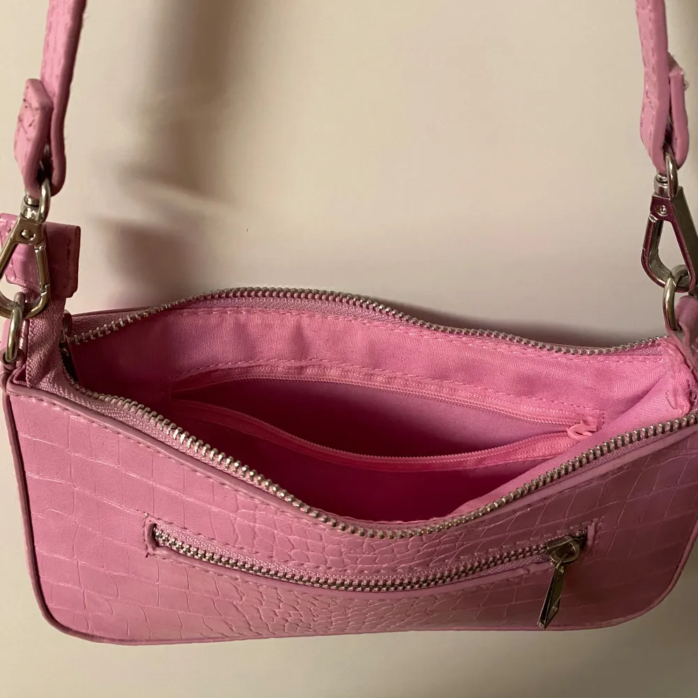 Snygg rosa axelväska i syntet med krokodilmönster. Väskan har en dragkedja upptill och en extra dragkedja på framsidan för småsaker. Perfekt för att ge din outfit en färgklick!. Laukut & Käsilaukut.