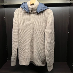 Beige stickad tröja från Massimo Dutti - Säljer en snygg beige stickad tröja från Massimo Dutti med dragkedja och en avtagbar blå huva. Perfekt för kyliga dagar med sin mysiga och stilrena design. Tröjan har långa ärmar och ribbade muddar för extra komfort.