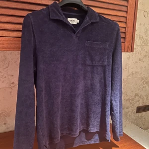 Mörkblå frottétröja från East West - Säljer en mörkblå frottétröja från East West. Tröjan har en klassisk krage och en bröstficka. Perfekt för en avslappnad stil. Den är långärmad och gjord i mjukt frottétyg.