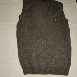Grå stickad väst från Lyle & Scott - Säljer en stilren grå stickad väst från Lyle & Scott med v-ringning och ett broderat gult emblem på bröstet. Perfekt för lager-på-lager under kyligare dagar. Klassisk design som passar till många olika stilar.