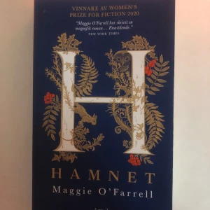Hamnet - Boken 'Hamnet' av Maggie O'Farrell, vinnare av Women's Prize for Fiction 2020. Omslaget är mörkblått med ett stort dekorativt 'H' i vitt och guld, omgiven av blommor och blad i guld och rött. En hyllad roman med citat från New York Times.