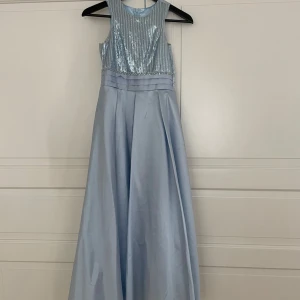 Ljusblå långklänning med paljetter - Elegant ljusblå långklänning med paljettöverdel och en stor rosett i ryggen. Klänningen har en ärmlös design och en vacker, flytande kjol. Perfekt för speciella tillfällen.