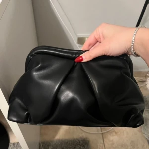 Svart handväska i syntet - Snygg svart handväska i syntetmaterial med en stilren design. Perfekt för att bära med sig det viktigaste. Enkel att matcha med olika outfits.